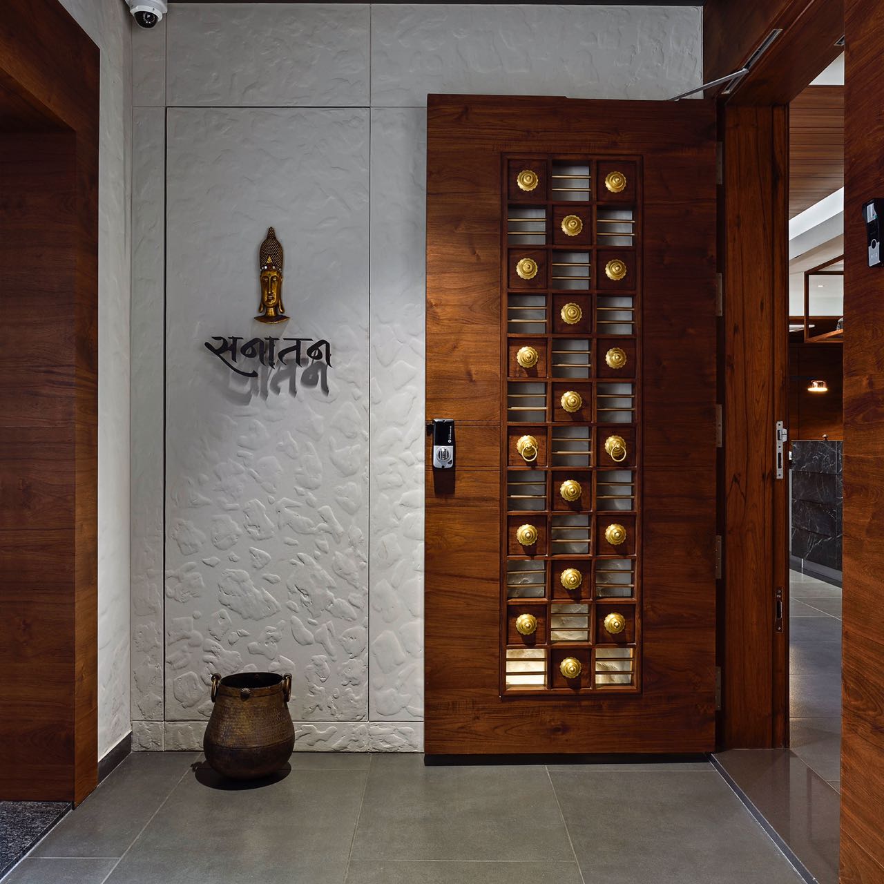 MA HOME entrance: carved teak door with brass studs beside a textured white wall and brass Buddha
